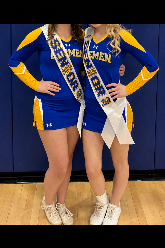 Two cheerleaders wearing matching royal blue and yellow Under Armour cheer outfits with white sashes that read 'SEMPER MEMORABLE' and 'MENEMEN', standing against a dark blue gym wall and wood floor.
