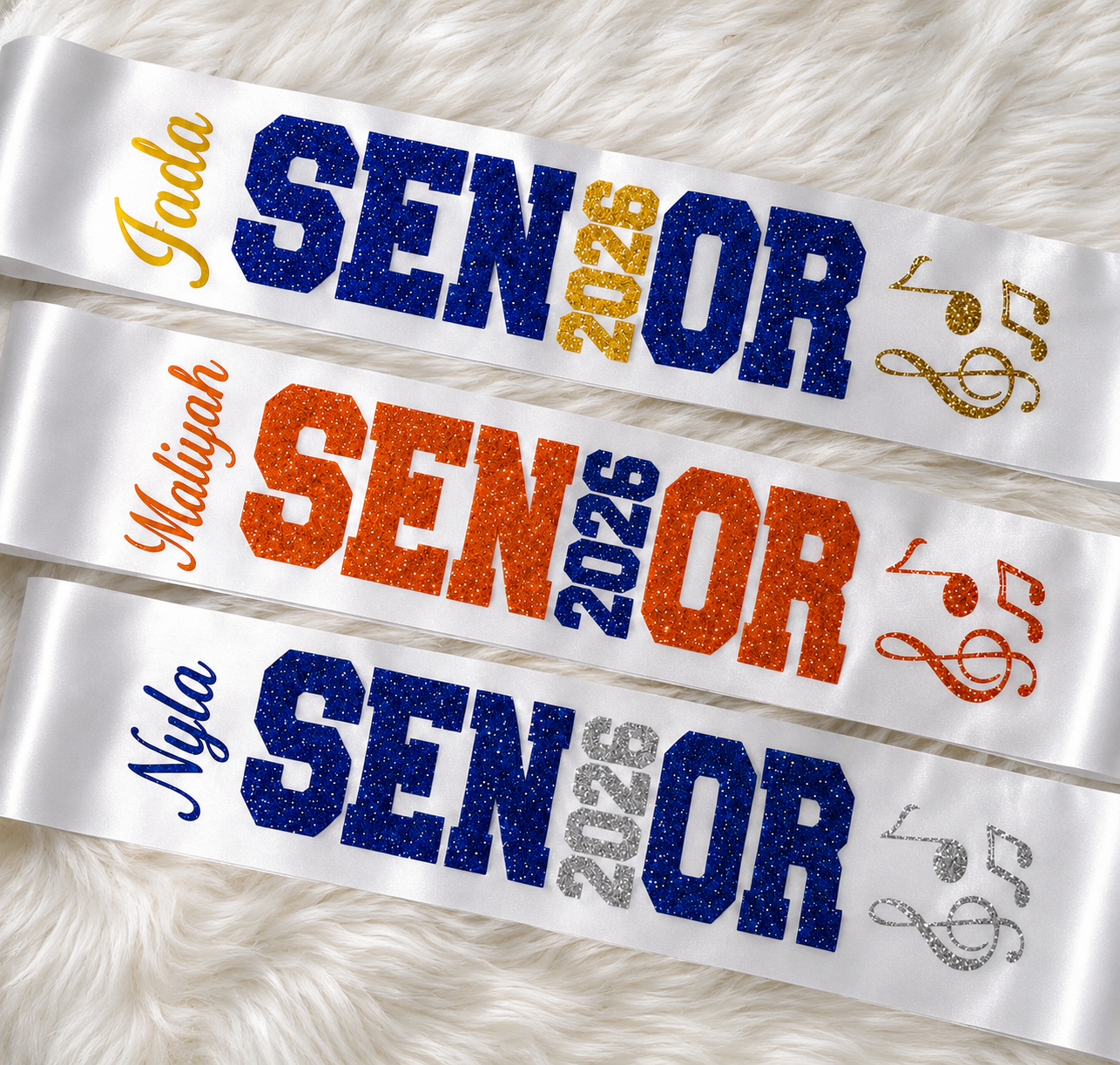 Three personalized sashes laid flat on a white faux-fur background. Each sash reads 'Jada SENIOR 2026', 'Maliyah SENIOR 2026', and 'Nyla SENIOR 2026' in glittery blue, orange, and silver lettering, with matching musical note accents.
