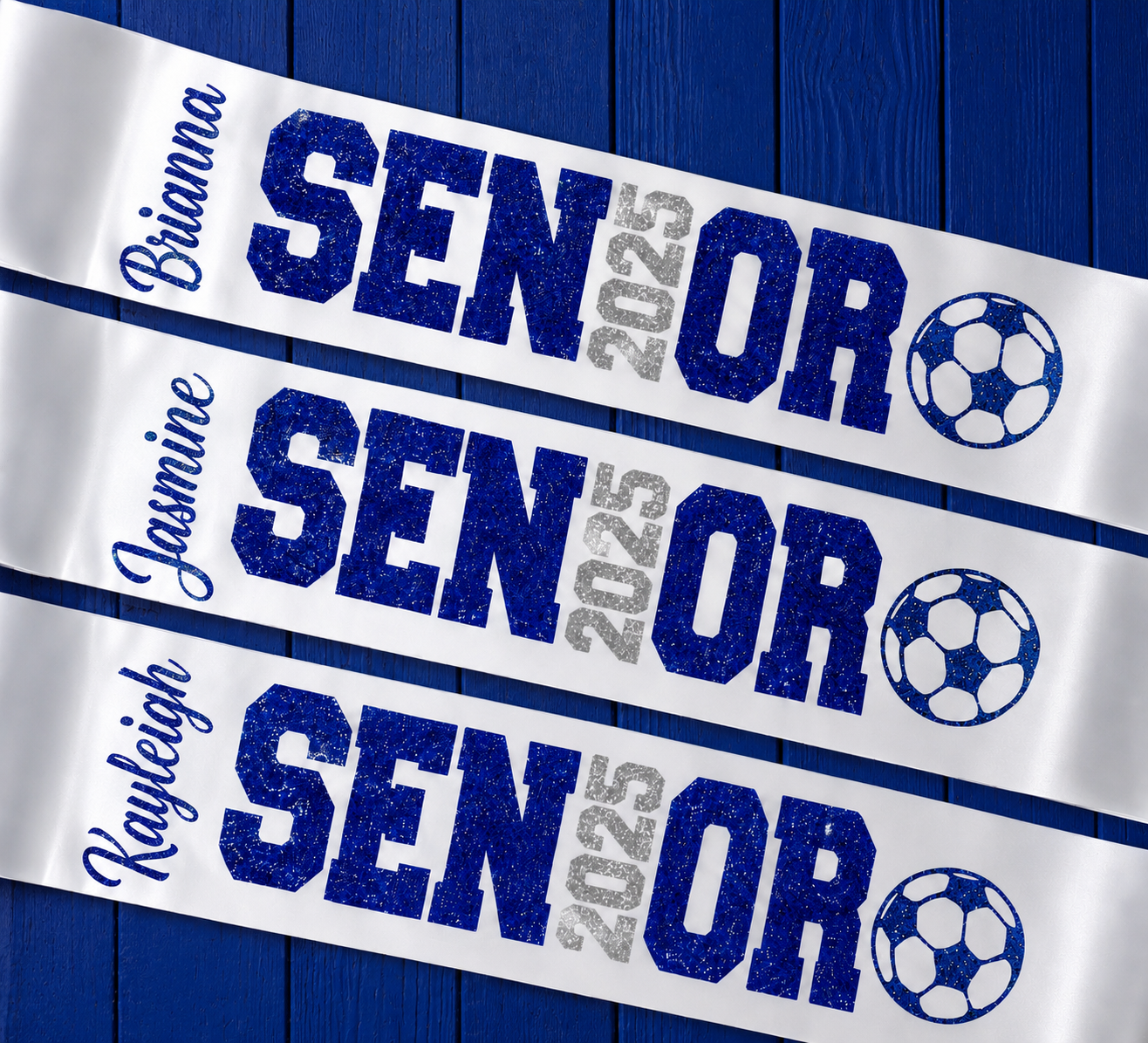 Three white personalized sashes laid diagonally on a dark blue wood background. Each sash has the name of a player in blue script, large blue glitter text reading SENIOR, gray glitter text reading 2025, and a soccer ball graphic.